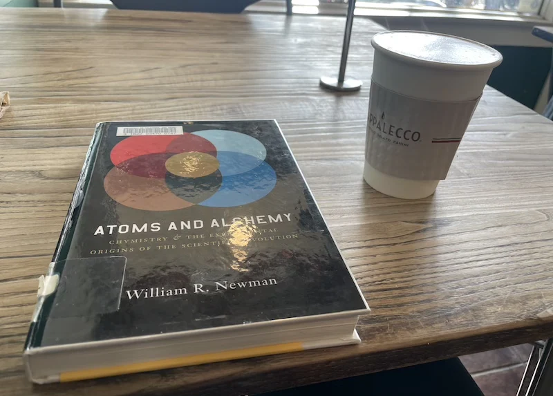 A picture of a book: Atoms and Alchemy sitting on a table beside a latte in the morning light.